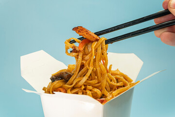 Udon stir-fry noodles with chicken meat and sesame in cardboard box on blue background close up. bright color.