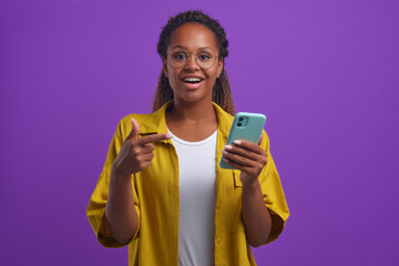 Young joyful inspired African American woman points to phone and sighs with smile after reading good news about career advancement or getting grant at university stands on plain lilac background