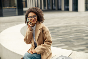 Wall Mural - Smiling afro american businesswoman talking phone while sitting on background of modern building 