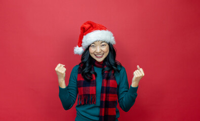 Wall Mural - Pretty smiling asian woman in warm christmas sweater and santa hat is having excitement on red background for season celebration concept with copy space