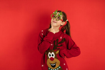 Sticker - A funny girl in a Christmas jumper and glasses, looks up and holds her hand near her face on a red background