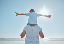 Family, Beach And Summer Vacation For Freedom With A Child On Shoulder Of Father With Hands Outstretched For Happiness Against Blue Sky. Man And Kid Son Together At Sea For Trust, Nature And Peace