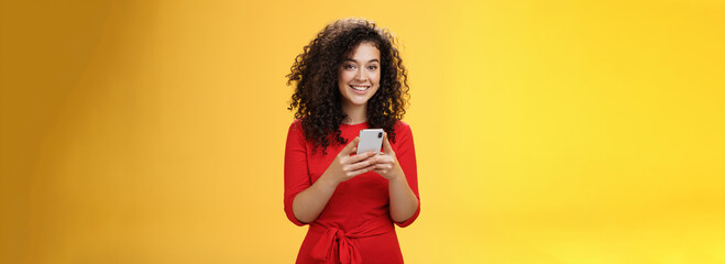 Girl messaging friend to tell all hot rumors holding mobile phone in hands smiling broadly and excited at camera as using smartphone browsing in net or edditing in app over yellow background