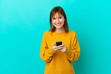 Wall Mural - Redhead girl isolated on blue background sending a message with the mobile