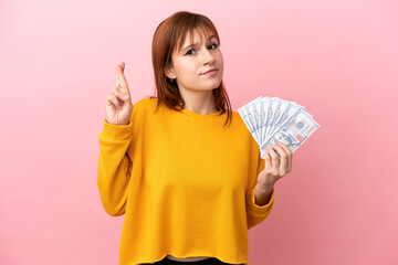 Wall Mural - Redhead girl taking a lot of money isolated on pink background with fingers crossing and wishing the best