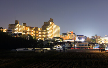 Well-lit apartment building over residential neighborhood next to small farm at night