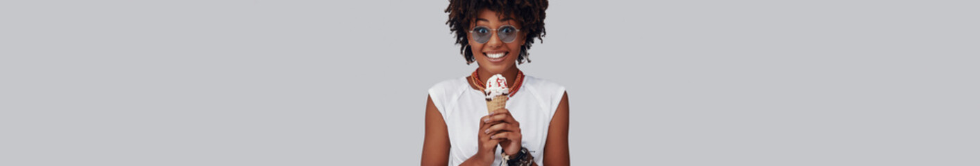 Poster - Happy young African woman enjoying ice cream while standing against grey background