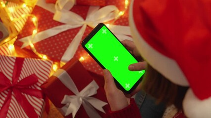 Poster - Human is holding a smartphone with a green screen on the background of Christmas tree branches with lights, red round toys and Christmas gift boxes. Mobile search, shopping, holiday sale.