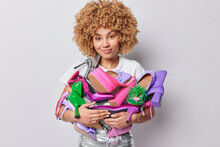 Horizontal Shot Of Good Looking Woman With Curly Hair Embraces Big Stack Of Fashionable Multicolored Shoes Prefers Wearing High Heels Poses Indoor Against Grey Studio Background Sells Her Footwear
