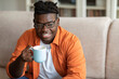 © Prostock-studio - Smiling black guy sitting on couch with mug of coffee