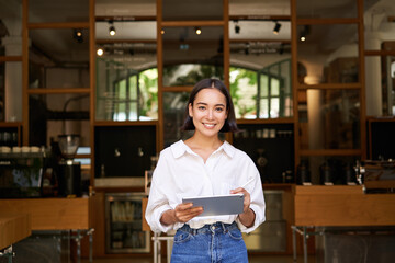 Portrait of asian woman, manager standing with tablet in front of cafe entrance, welcomes guests