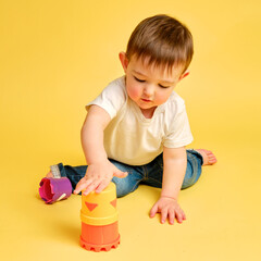 Wall Mural - Toddler baby is playing logical educational games with a sandbox mold on a studio yellow background. Happy child playing with an educational toy bucket, learning logic. Kid aged one year four months