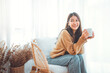 © oatawa - Happy asian woman relaxing drinking hot coffee or tea in holiday morning vacation on armchair at home, Cosy scene, Smiling pretty woman drinking hot tea in autumn winter. copy space.