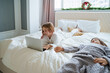 © Westend61 - Girl using laptop lying on bed by brother at home
