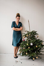 Woman Standing With Arms Crossed Looking At Broken Bauble By Christmas Tree In Front Of White Wall
