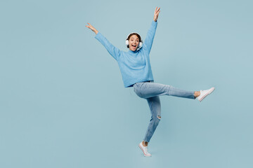 Full body young happy woman wear knitted sweater headphones listen to music raise up hands dance have fun isolated on plain pastel light blue cyan background studio portrait. People lifestyle concept.