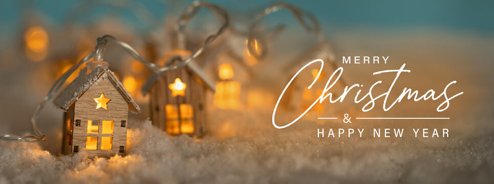 Fototapete - Christmas Greeting Card, English text. Abstract Christmas Winter Panorama with Wooden Houses Christmas String Lights in Cold Snow Landscape and Glowing Golden Lights in Background. Panorama, Banner. 
