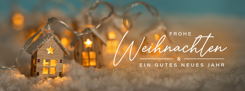 Fototapete - Christmas Greeting Card, German text. Abstract Christmas Winter Panorama with Wooden Houses Christmas String Lights in Cold Snow Landscape and Glowing Golden Lights in Background. Panorama, Banner. 