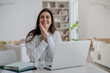 © Iona - Excited brunette caucasian young adult woman in white shirt sitting at desk with laptop leans on folded palms looks up dreamily toothy smiles. Successful businesswoman happy after distant meeting.
