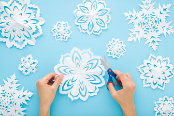 Young adult woman hands cutting different white snowflake shapes from paper on light blue table background. Pastel color. Making decoration elements for winter festive. Closeup. Top down view.