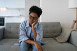 Tired exhausted African American woman sits on sofa in living room leaning on arm and looking into distance. Thoughtful young black in glasses and casual clothes is resting after hard day. Boredom.