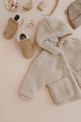 Wall Mural - Stylish elegant baby clothes, accessories and toys on neutral beige background. Warm winter fleece jacket, suede booties, hat, pacifier, toys. Neutral pastel beige colour. Flat lay, top view