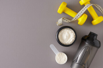 Fitness diet concept. Top view composition made of protein powder in jar with shaker, dumbbells and tape measure on grey background with copy space. Sport food supplement idea.