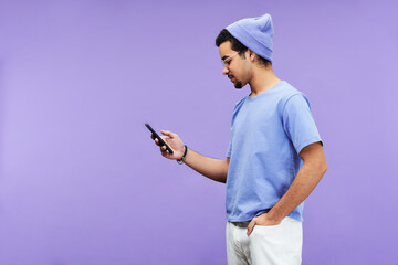 Aufkleber - Side view of serious guy in blue t-shirt and beanie hat texting in mobile phone or looking through online information on violet background