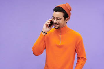 Aufkleber - Happy guy in eyeglasses, orange pullover and beanie hat talking on mobile phone in front of camera against violet background