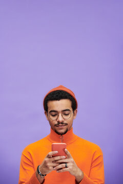 Young serious man in orange casualwear and eyeglasses texting in smartphone while standing against lavender background