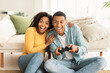 Happy excited young african american wife and husband sit on floor with joystick, play in online game