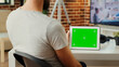 © DC Studio - Young man holding modern device with horizontal greenscreen template in living room, sitting at desk. Using isolated chroma key display with copy space and mock up backgroung, screen app.