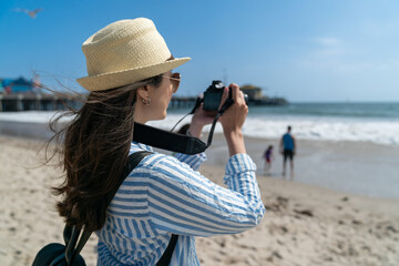 Wall Mural - back view of young asian Japanese woman photographer wearing hat taking photos of beautiful ocean waves with slr camera in breeze