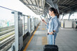 Asian woman commuter standing on the subway platform waiting to go inside train. She is using a smart phone with a suitcase.