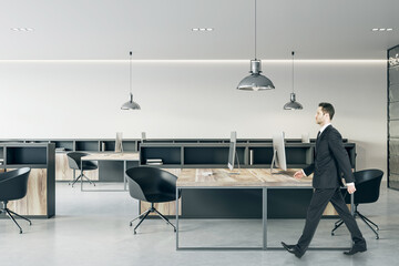 Aufkleber - Businessman in black suit walking through wooden work table with modern computers in open space modern office with dark stylish furniture and light wall background