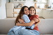 © DimaBerlin - Young woman loving mother hugging embracing her happy adopted daughter at home, expressing love to teenage child, joyful mom and child sitting together on couch and cuddling. Family reunion concept