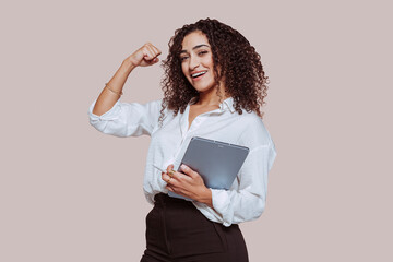 Portrait of a young cheerful arabic businesswoman, showing strong bicep muscle, works using digital tablet isolated over beige background at studio. Work, communication, technology concept.