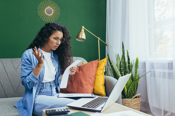 Frustrated and sad woman at home paying bills and paying mortgage, hispanic woman has no money for paperwork using laptop and calculator to calculate household budget sitting on sofa.