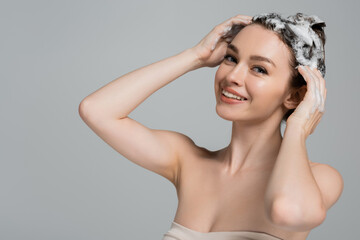 cheerful young woman washing wet and foamy hair isolated on grey