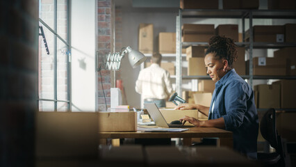 Wall Mural - Black Female Warehouse Inventory Manager Works on Laptop Computer while Sitting at Her Desk. In the Background Employee Working in the Room Full of Cardboard Box Parcels Ready For Shipping.