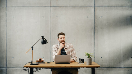 Poster - Professional Creative Man Sitting at His Desk in Office Working on a Laptop Computer, Working, Thinking, Solving Problems and Smiling at Success. Stylish Office Space Design Perfect for Productivity