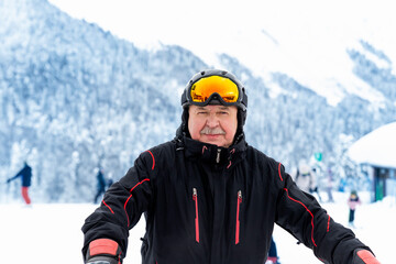 Active senior experienced mature old person in ski helmet, goggles and black suit stand against scenic panorama peak enjoy cold extreme sport activities. Man on mountains forest winter background