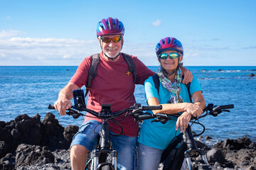 Cheerful Caucasian senior couple posing for a portrait on the beach riding on electric bicycles. Authentic retirement living and healthy lifestyle concept. Horizon over the sea