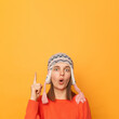 © sementsova321 - Indoor shot of shocked woman wearing funny earflap hat and orange casual jumper standing isolated over yellow background, pointing up with index finger, showing advertisement area.