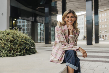 Smiling caucasian young lady spends time outdoors near business center. Blonde woman wears casual clothes in spring. Concept of enjoying moment