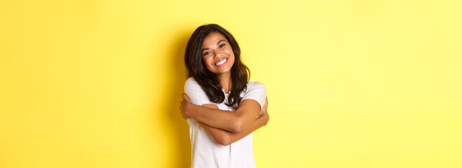 Portrait of cheerful african american woman, hugging herself and smiling pleased, standing over yellow background