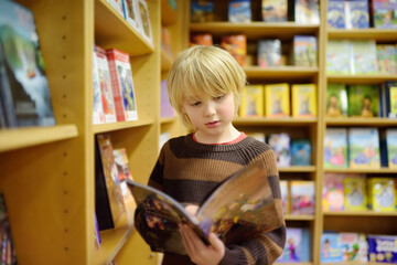 A preteen boy leafing through a book while standing at a bookshelf in a school library or bookstore.