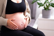 Unrecognizable pregnant woman sitting on bed caressing naked baby bump, belly, wearing black jeans and top indoors at home. Happy,natural pregnancy, care concept .