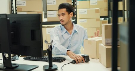 Wall Mural - Happy Asian man wear formal uniform shirt sit in front of desk use computer and barcode machine scanner record customer online data in cardboard box delivery at warehouse. Startup business concept.