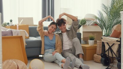 Wall Mural - Happy Asian couple sit on floor in front of sofa couch smile looking at camera making heart sign in living room at new house. Husband and wife moving new house, Apartment Rent And Ownership Concept.